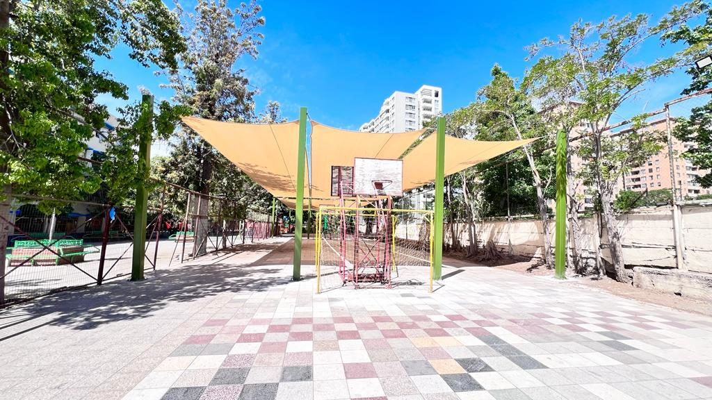 Vela de sombra sobre una cancha de básquetbol escolar rodeada de árboles