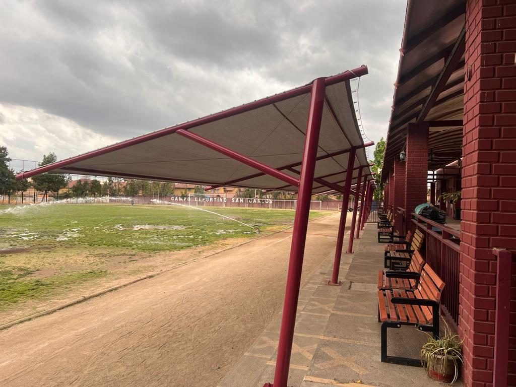 Graderías techadas con estructura metálica burdeo y bancas frente a una cancha de fútbol