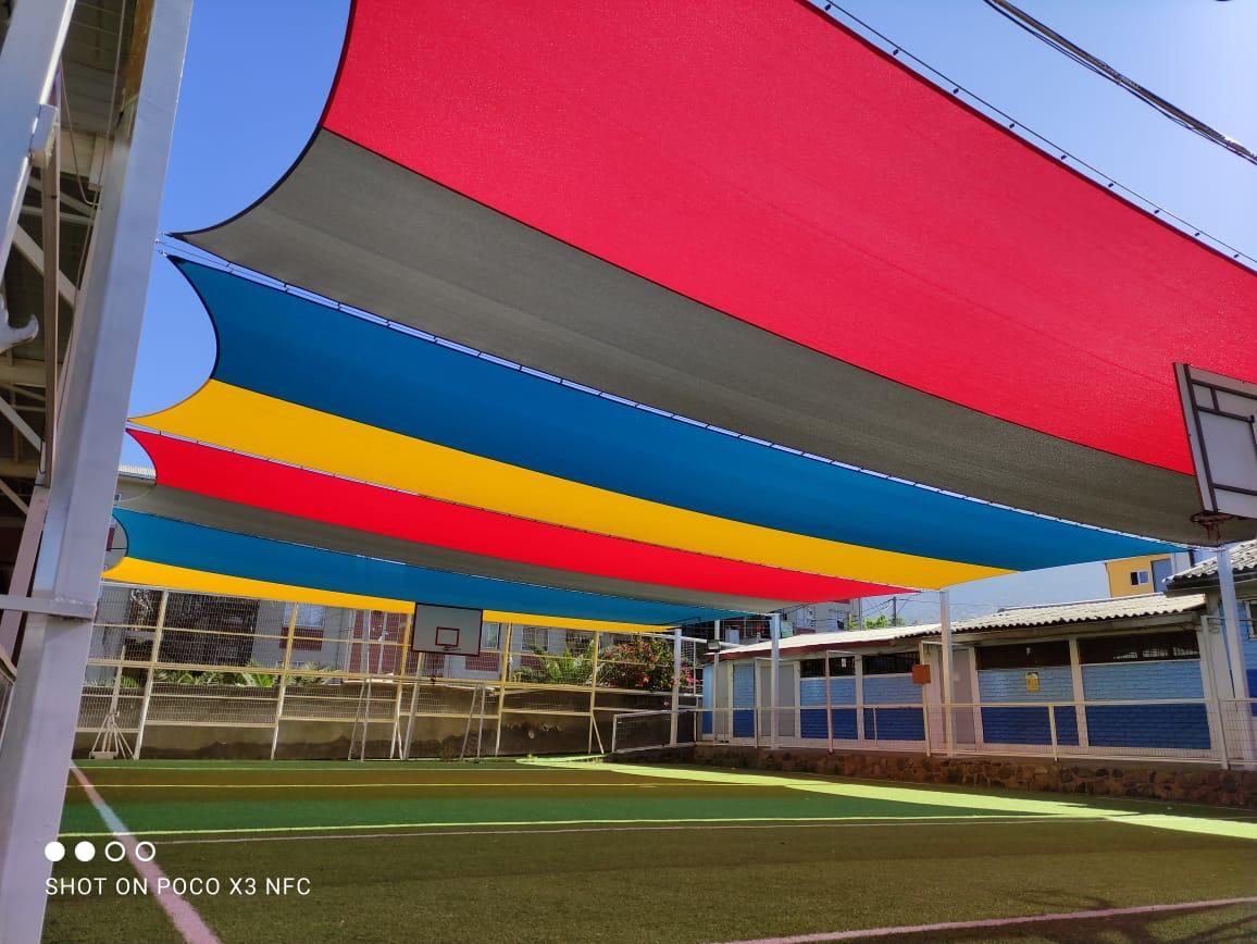 Velas de sombra tensadas en rojo, azul, amarillo y gris sobre una cancha de futsal con pasto sintético